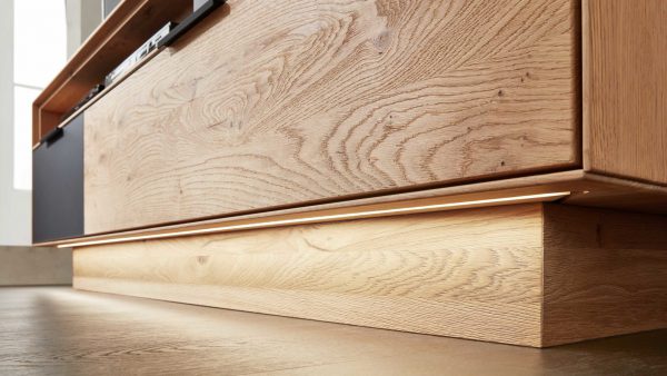 Nahaufnahme eines modernen Holzschranks aus der Interliving Wohnzimmer Serie 2005 mit Unterbaubeleuchtung 195 053, einem darunter installierten LED-Lichtband, das einen warmen Lichtschein auf den Naturholzboden wirft.
