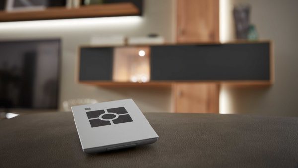 Die Interliving Wohnzimmer Serie 2005 - Funkdimmer 101 004, eine kompakte Fernbedienung mit schwarzem Quadrat und kreisförmigem Design, ruht auf dunklem Leder, während sanft beleuchtete Regale und Schränke im Hintergrund verschwimmen.