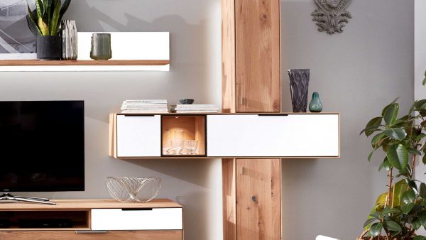 In einem modernen Wohnzimmer steht die Interliving Wohnzimmer Serie 2005 - Quer-Vitrine 117 821 in hellem Holz und Weiß, mit dekorativen Vasen, Büchern, einer Pflanze, einem Fernseher auf einem Schrank und einer Glasschale darunter. Das Sonnenlicht füllt den Raum.