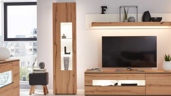 Modernes Wohnzimmer mit der Interliving Wohnzimmer Serie 2103 - Vitrine 560720, einem Hochschrank mit Glasböden, einem Fernseher auf einem niedrigen Ständer, ausgestellten Dekorationsgegenständen und großen Fenstern für natürliches Licht.