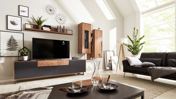 In einem modernen Wohnzimmer steht die Interliving Wohnzimmer Serie 2106 - Wohnwand 620001, gepaart mit einem schwarzen Sofa, dekorativen Pflanzen, gerahmten Kunstwerken, einem Couchtisch zum Essen und großen Fenstern, die den Raum mit natürlichem Licht durchfluten.