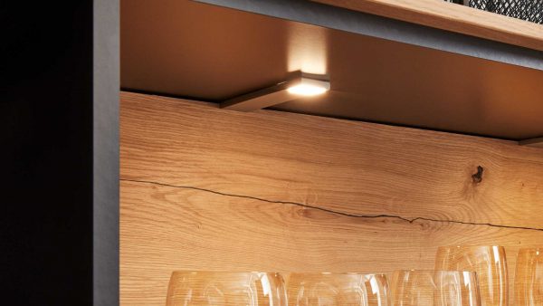 Eine Nahaufnahme eines Holzregals mit dem Interliving Wohnzimmer Serie 2106 - LED-Unterbauspot 22-86, der klare Glasbecher darunter beleuchtet. Im Hintergrund sind die natürliche Holzmaserung und ein markanter Riss zu sehen.