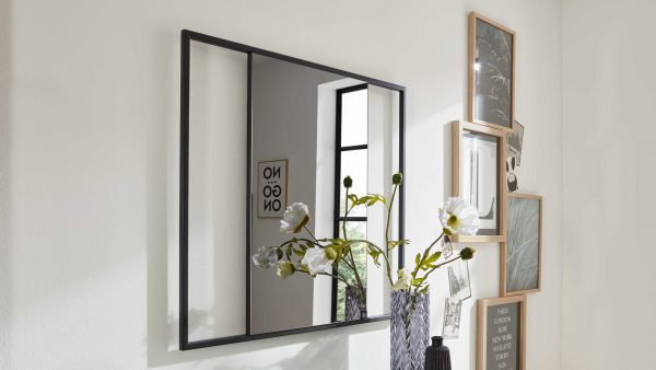 Der Interliving Garderoben Serie 6009 - Wandspiegel 661921, ein quadratischer, schwarz gerahmter Spiegel, reflektiert eine Wand, ein Fenster und einen NO GO Druck. Davor steht eine Vase mit weißen Blumen, an der Wand daneben hängen gerahmte Bilder.