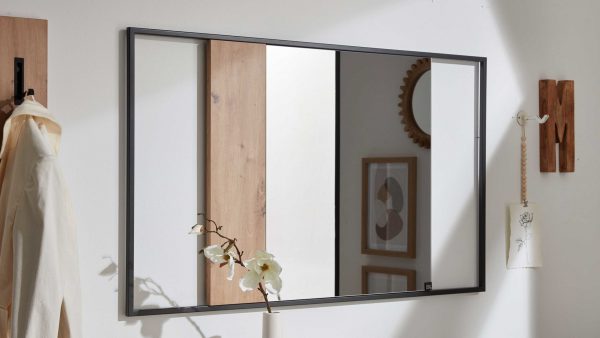 Der Interliving Garderoben Serie 6009 - Wandspiegel 661911, ein rechteckiger, schwarz gerahmter Spiegel an einer weißen Wand, reflektiert Wandkunst, einen runden Spiegel und eine Holzplatte. In der Nähe befinden sich eine Orchidee und eine helle Jacke.