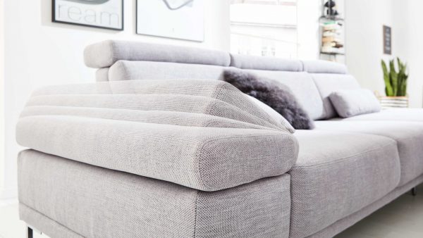 Nahaufnahme des Interliving Sofas Serie 4251 mit verstellbarer Armlehne in hellgrauem Stoff in einem hellen Wohnzimmer. Im Hintergrund sind ein dunkles Kissen, gerahmte Wandkunst und eine grüne Topfpflanze zu sehen.