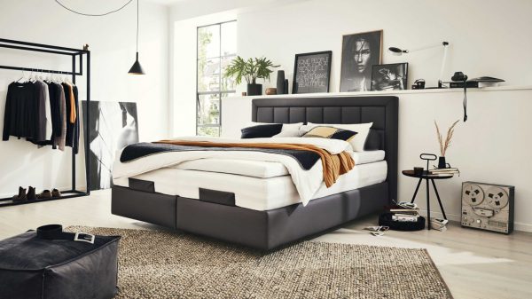 Modernes Schlafzimmer mit dem Interliving Boxspringbett Serie 1410, Lagenbettwäsche, gerahmter Kunst auf einem Regal, Kleiderständer, Sonnenfenster, Pflanze, Teppich und einem alten Kassettenrekorder für eine minimalistische Einrichtung.
