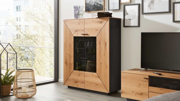 Ein modernes Wohnzimmer mit einem großen Fenster, in dem die Interliving Wohnzimmer Serie 2022 - Highboard HB2 Vitrine mit Glastür, ein Fernseher auf einem Holzständer, ein Weidenkorb, eine Pflanze und gerahmte Kunst an der Wand stehen.
