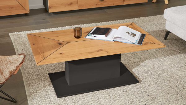 Der Interliving Wohnzimmer Serie 2022 - Couchtisch CTV330-110, ein moderner Couchtisch aus Holz mit schwarzem, rechteckigem Sockel, steht auf einem hellen Teppich mit einem offenen und einem geschlossenen Buch sowie einer dunklen Glastasse; daneben stehen ein Sofa und andere Holzmöbel.