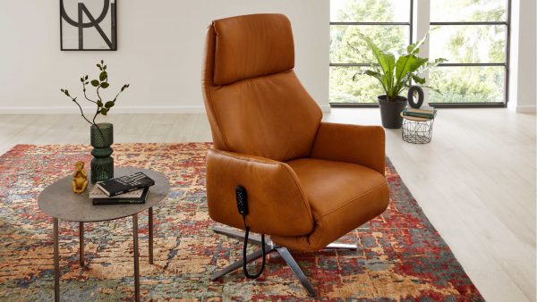 In einem modernen Wohnzimmer steht der Interliving Sessel Serie 4520 - Sessel 56U46, ein hellbrauner Ledersessel, auf einem farbenfrohen Teppich vor großen, begrünten Fenstern. Ein runder Tisch mit einer Vase, Büchern und Gläsern sowie einer Blattpflanze steht neben dem Fenster.