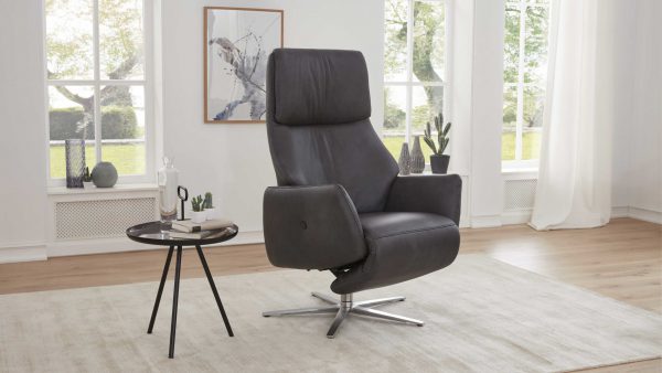 Der Interliving Sessel Serie 4520 - Sessel 26U44, ein moderner schwarzer Ledersessel mit hoher Rückenlehne, steht auf einem hellen Teppich in einem hellen Raum neben einem runden Beistelltisch mit Büchern und Dekoration, gerahmter Kunst und großen Fenstern im Hintergrund.