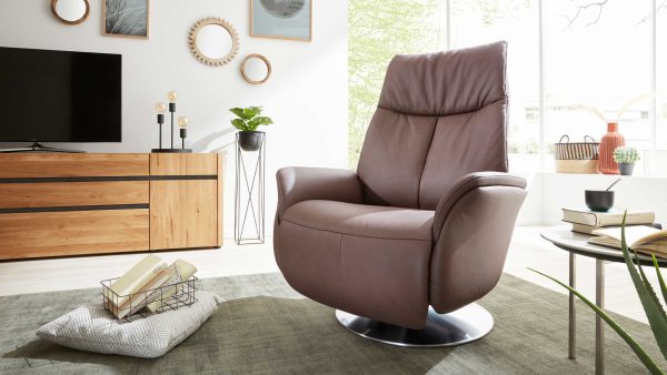 Ein modernes Wohnzimmer mit dem braunen Ledersessel Sessel Serie 4550 - Sessel 37O44 von Interliving auf einem runden Sockel, gepaart mit einem hölzernen TV-Schrank, einem runden Couchtisch mit Büchern, Wandkunst, hellem Holzboden und einem grünen Teppich.