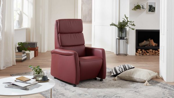 Ein modernes Wohnzimmer mit dem OrthoSedis Relaxsessel OS-HU1043 mit Aufstehhilfe mit Federkern in kastanienbraun, einem runden weißen Couchtisch mit Büchern und Pflanzen, einem Kamin mit gestapelten Holzscheiten, Kissen auf einem hellen Teppich und Topfpflanzen an hellen Fenstern.