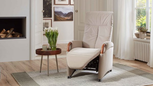 Der OrthoSedis OS-HU1017 motorisierte Relaxsessel in Beige mit Holzarmlehnen steht auf einem hellen Teppich neben einem runden Beistelltisch mit weißen Tulpen in einem hellen, gemütlichen Wohnzimmer mit Kunst und großen Fenstern.