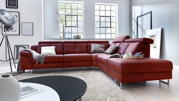 In einem modernen Wohnzimmer steht das Interliving Sofa Serie 4050 - Ecksofa in Rot mit grauen und gemusterten Kissen, einer gemütlichen Decke, Wandkunst, großen Fenstern und einem grauen Teppich. Natürliches Licht durchflutet den Raum und sorgt für eine helle, einladende Atmosphäre.