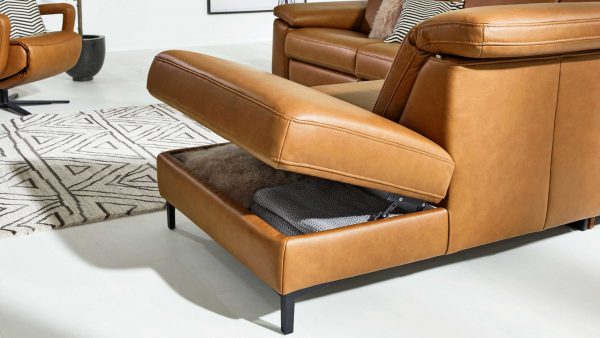 Das Interliving Sofa Serie 4054 - Aufklappfunktion F ist mit hellbraunem Leder ausgestattet und verfügt über einen hochklappbaren Sitz, der Stauraum mit einer gefalteten Decke bietet. Im Hintergrund sind ein gemusterter Teppich und abgestimmte Möbel zu sehen.