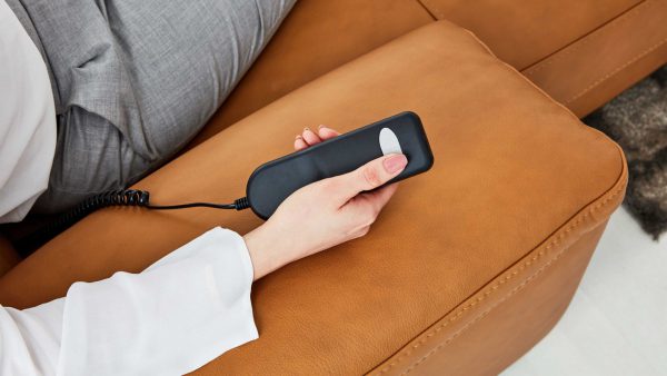 Eine Person in grauer Hose und weißem Hemd liegt auf dem Interliving Sofa Serie 4054 und hält die Handschalter HS-Fernbedienung mit weißem Knopf.
