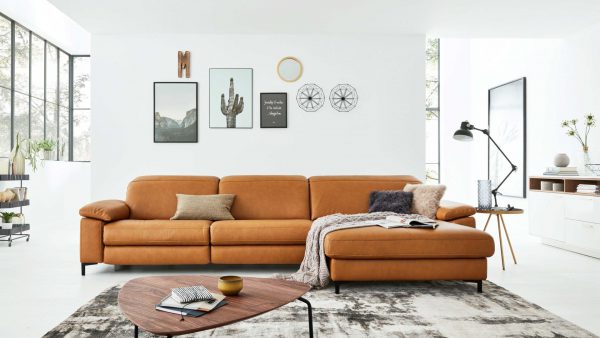 In einem modernen Wohnzimmer steht das Interliving Sofa Serie 4054 - Ecksofa, das mit verschiedenen Kissen, einer Decke und Zeitschriften dekoriert ist. Dahinter befinden sich gerahmte Bilder und dekorative Wandkunst an einer weißen Wand, vor der ein Couchtisch aus Holz steht.