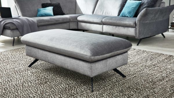 Ein modernes Wohnzimmer mit dem Interliving Sofa Serie 4056 XXL-Funktionshocker, einer großen grauen Ottomane auf einem strukturierten Teppich, gepaart mit einer passenden grauen Couchgarnitur und blauen Akzentkissen im Hintergrund.