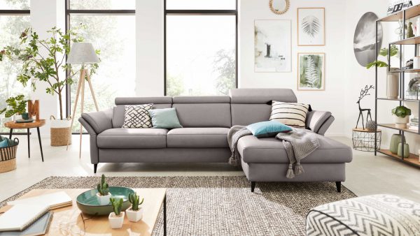 Modernes Wohnzimmer mit dem Interliving Sofa Serie 4055 - Ecksofa, das mit dekorativen Kissen, Zimmerpflanzen, gerahmter Wandkunst, einer Stehlampe und einem runden Couchtisch auf einem gemusterten Teppich vor hohen, sonnenbeschienenen Fenstern eingerichtet ist.