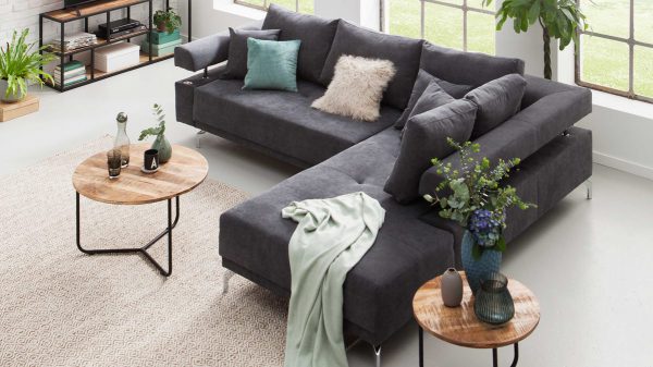 Modernes Wohnzimmer mit dem KAWOO Funktions-Ecksofa bzw. Polsterecke Dream in Dunkelgrau, verschiedenen Kissen, einer hellen Decke, zwei runden Couchtischen aus Holz, Topfpflanzen und großen Fenstern, die das Tageslicht hereinlassen.