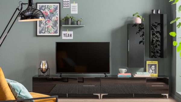 Ein modernes Wohnzimmer mit der MAJA MÖBEL Wohnkombination Shino, mit einem schwarzen Fernseher auf einem schlichten dunklen Schrank, Wandregalen für Dekoration, gerahmter Kunst, Büchern, einer Lampe und einer Tasse mit Untertasse. Die Wände sind in einem sanften Grün-Grau gestrichen.