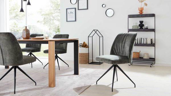 Modernes Esszimmer mit Interliving Esszimmer Serie 5110 - Polsterstuhl CO810 aus grünem Samt, einem Holztisch, minimalistischen schwarzen Regalen mit Büchern und Dekoration, gerahmter Wandkunst und großen Fenstern, die den Raum mit natürlichem Licht füllen.
