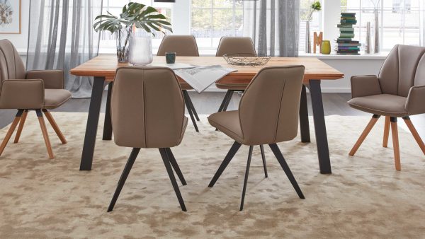 Ein modernes Esszimmer mit einem Holztisch, sechs Stühlen der Interliving Esszimmer Serie 5102 - Polsterstuhl V321, einem beigen Teppich und einem dekorativen Mittelstück. Große Fenster mit transparenten Vorhängen lassen natürliches Licht herein.