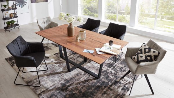 Ein modernes Esszimmer mit der Interliving Esszimmer Serie 5107 - Esstisch 8473, fünf gemischten schwarzen und beigen Stühlen, dekorativen Vasen mit Blumen, Büchern und Papieren auf dem Tisch, großen Fenstern und einem gemusterten Teppich auf hellem Holzfußboden.