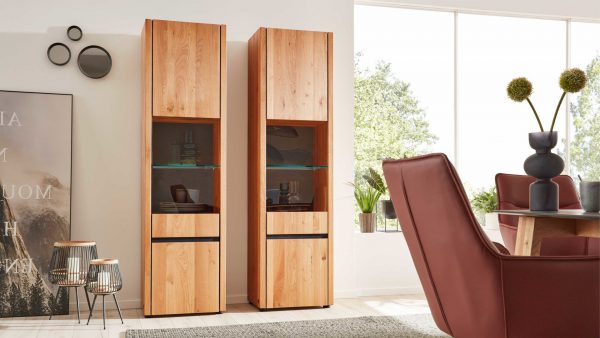 Zwei Interliving Esszimmer Serie 5603 - Vitrine 3774 mit Glasböden stehen nebeneinander in einem modernen Wohnzimmer, neben einem roten Stuhl und einem Esstisch. Das Sonnenlicht fällt durch große Fenster und hebt Vasen und Wandkunstakzente hervor.