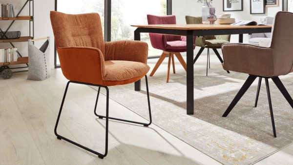 In einem modernen Esszimmer stehen die Stühle der Interliving Esszimmer Serie 5111 - Schalensessel 9032-04 um einen Holztisch, der auf einem hellen Holzfußboden steht. Das Sonnenlicht fällt durch große Fenster in den Raum, während im Hintergrund Bücher und Dekoration in Regalen stehen.