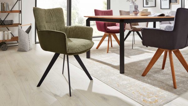 Ein modernes Esszimmer mit hellem Holzfußboden, einem Holztisch und Interliving Esszimmer Serie 5111 - Schalensessel 9022-04 Stühlen in Grün, Lila und Rosa auf einem gemusterten Teppich. Durch die großen Fenster im Hintergrund fällt natürliches Licht ein.
