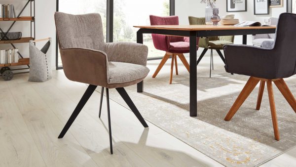 In einem modernen Essbereich stehen die Stühle der Interliving Esszimmer Serie 5111 - Schalensessel 9022-04 in verschiedenen Farben um einen Holztisch herum, der auf einem hellen Teppich und einem hellen Boden steht, mit großen Fenstern und Bücherregalen dahinter.