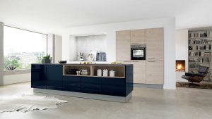 Moderne Küche mit einer Insel in glänzendem Marineblau, Holzschränken, eingebautem Ofen und offenen Regalen. Große Fenster sorgen für natürliches Licht, und im angrenzenden Raum ist ein gemütlicher Kamin mit einem Sessel zu sehen.