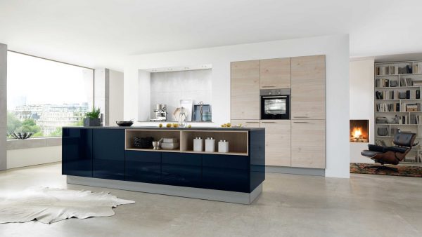 Moderne Küche mit einer Insel in glänzendem Marineblau, Holzschränken, eingebautem Ofen und offenen Regalen. Große Fenster sorgen für natürliches Licht, und im angrenzenden Raum ist ein gemütlicher Kamin mit einem Sessel zu sehen.