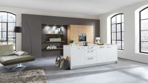 Moderne Küche mit großen Fenstern, einer weißen Insel mit Schubladen, Topfpflanzen, Geräten und Küchenutensilien. Holzschränke, offene Regale und ein Loungesessel vervollständigen den hellen, stilvollen Raum.