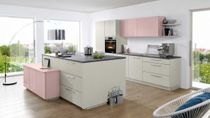 Moderne Küche mit weißen und hellrosa Schränken, grauen Arbeitsplatten, eingebautem Ofen, Herd und Kaffeemaschine. Große Fenster und Glastüren lassen natürliches Licht herein, mit Blick auf das Grün im Freien. Holzboden und minimalistisches Dekor.