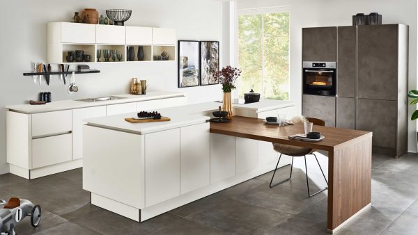 Moderne Küche mit weißen Schränken, grauen Geräten, einem eingebauten Backofen und einem an der Insel angebrachten Esstisch aus Holz. Zur Ausstattung gehören Regale mit Geschirr, Pflanzen und große Fenster, die natürliches Licht hereinlassen.