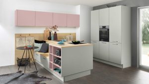 Moderne Küche mit hellgrauen Schränken, rosafarbenen Oberschränken, einer zentralen Insel mit offenen Regalen, Arbeitsflächen aus Holz und minimalem Dekor. Natürliches Licht fällt durch eine Tür ein, und auf dem dunklen Holzboden liegt ein Kuhfellteppich.