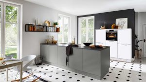Moderne Küche mit grauen Schränken, einer Insel mit schwarzer Arbeitsplatte, weißen Schränken mit Backofen, offenen Regalen mit Dekoration und großen Fenstern, die natürliches Licht hereinlassen. Der Boden hat ein schwarz-weißes Kachelmuster.