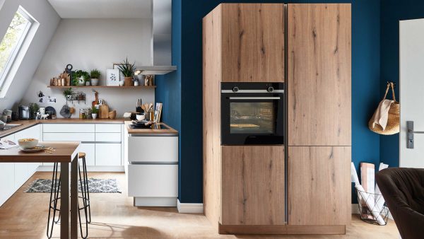 Moderne Küche mit weißen Schränken, Holzarbeitsplatten und einem eingebauten Ofen in hohen Holzschränken. Der Raum verfügt über blaue Akzentwände, Pflanzen und eine schräge Decke mit einem großen Fenster, das natürliches Licht hereinlässt.