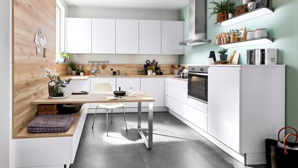 Moderne Küche mit weißen Schränken, eingebautem Backofen und offenen Regalen mit Pflanzen und Küchenutensilien. Ein kleiner Esstisch mit einem Stuhl steht an einer holzgetäfelten Wand, und das Sonnenlicht erhellt den aufgeräumten Raum.