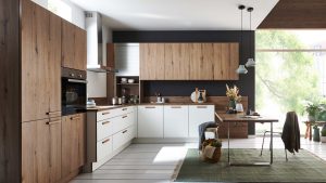 Moderne Küche mit Holz- und weißen Schränken, Einbaubackofen, Arbeitsplatten und offenen Regalen. Ein Esstisch mit Stühlen steht auf einem grünen Teppich neben großen Fenstern mit Blick auf Bäume und natürliches Licht.