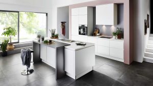 Moderne Küche mit weißen Schränken, großen Fenstern, Zimmerpflanzen und einer zentralen Insel mit Spüle und Barhocker; natürliches Licht erfüllt den Raum und schafft eine klare und einladende Atmosphäre.