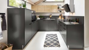 Moderne Küche mit matten dunkelgrauen Schränken, hellen Holzoberschränken, eingebautem Backofen, schwarzer Arbeitsplatte und einem geometrischen Teppich auf weißem Boden. Durch die oberen Fenster fällt helles Tageslicht ein und schafft eine gemütliche Atmosphäre.
