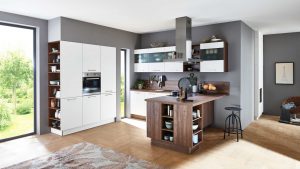 Moderne Küche mit weißen Schränken, Holzakzenten und offenen Regalen. Auf der rechten Seite befindet sich eine kleine Insel mit Hockern. Große Fenster lassen natürliches Licht herein, und die verschiedenen Küchenutensilien sind ordentlich auf Theken und Regalen angeordnet.