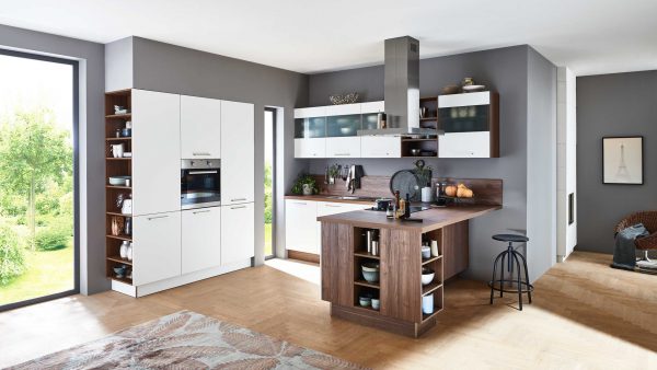 Moderne Küche mit weißen Schränken, Holzakzenten und offenen Regalen. Auf der rechten Seite befindet sich eine kleine Insel mit Hockern. Große Fenster lassen natürliches Licht herein, und die verschiedenen Küchenutensilien sind ordentlich auf Theken und Regalen angeordnet.