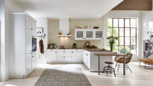 Helle, moderne Küche mit weißen Schränken, Einbaubackofen, einer zentralen Insel mit Barhockern, großen Fenstern, die natürliches Licht hereinlassen, Pflanzen und gemütlicher Einrichtung. Helle Holzböden und ein strukturierter Teppich vervollständigen den einladenden Raum.