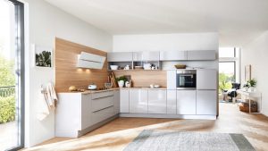 Moderne Küche mit glänzend weißen Schränken, hölzerner Aufkantung, eingebautem Ofen und offenen Regalen. Ein großes Fenster lässt natürliches Licht herein, und zur Ausstattung gehören Pflanzen, Geschirr und ein gemütlicher Teppich auf dem Holzboden.
