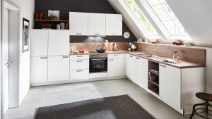 Moderne weiße L-förmige Küche unter einer schrägen Decke mit großen Fenstern, hölzernen Arbeitsplatten, eingebautem Ofen, Spüle, Regalen mit Geschirr und einem schwarzen Teppich auf dem Boden. Natürliches Licht erhellt den Raum.