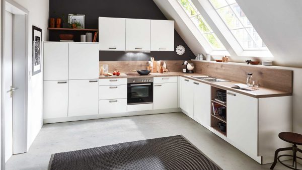 Moderne weiße L-förmige Küche unter einer schrägen Decke mit großen Fenstern, hölzernen Arbeitsplatten, eingebautem Ofen, Spüle, Regalen mit Geschirr und einem schwarzen Teppich auf dem Boden. Natürliches Licht erhellt den Raum.